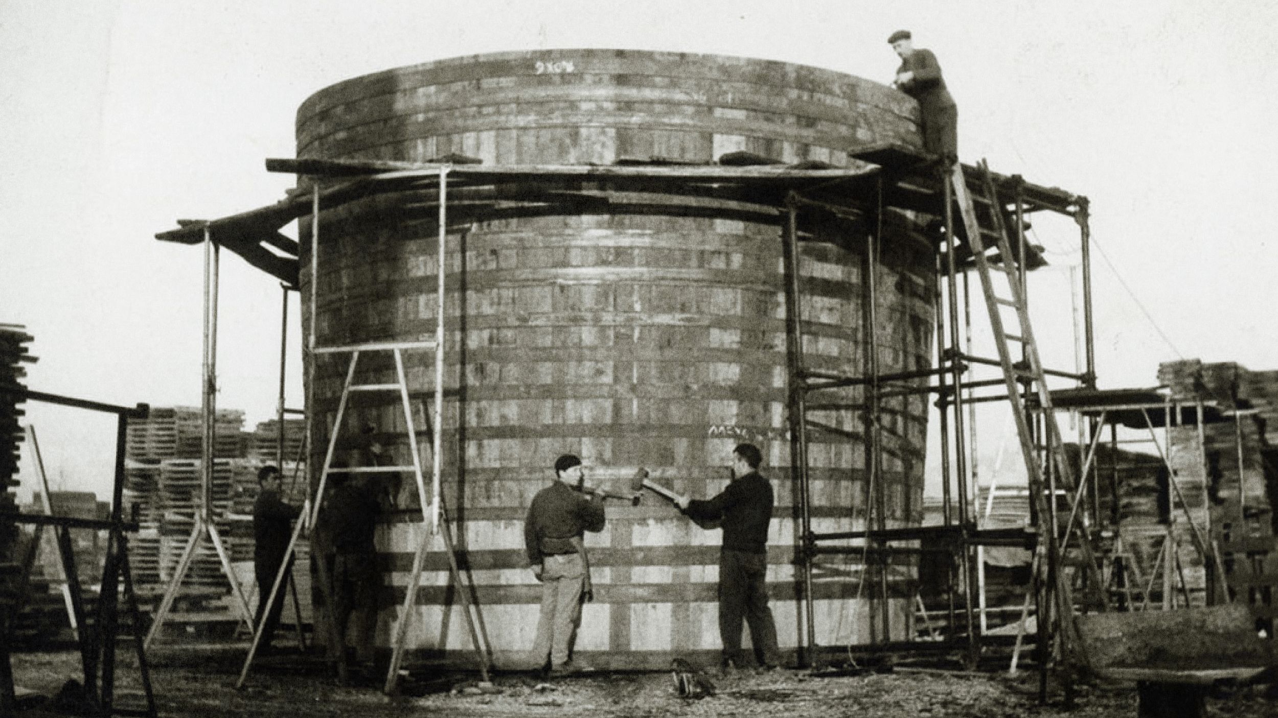 Four workers assemble a large wooden tank using scaffolding and ladders at an outdoor construction site, with stacks of materials in the background.