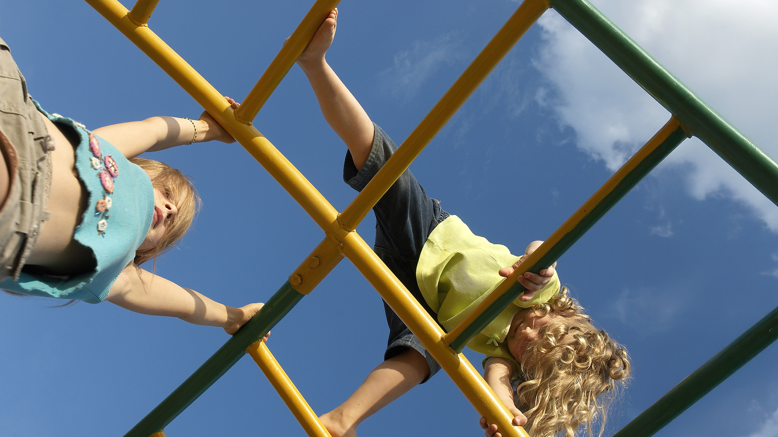 Two children are climbing on a yellow and green jungle gym, viewed from below against a blue sky with some clouds.
