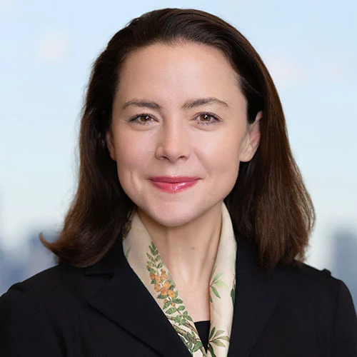 Woman with straight brown hair wearing a dark blazer and a light-colored scarf, smiling at the camera with a blurred cityscape background.