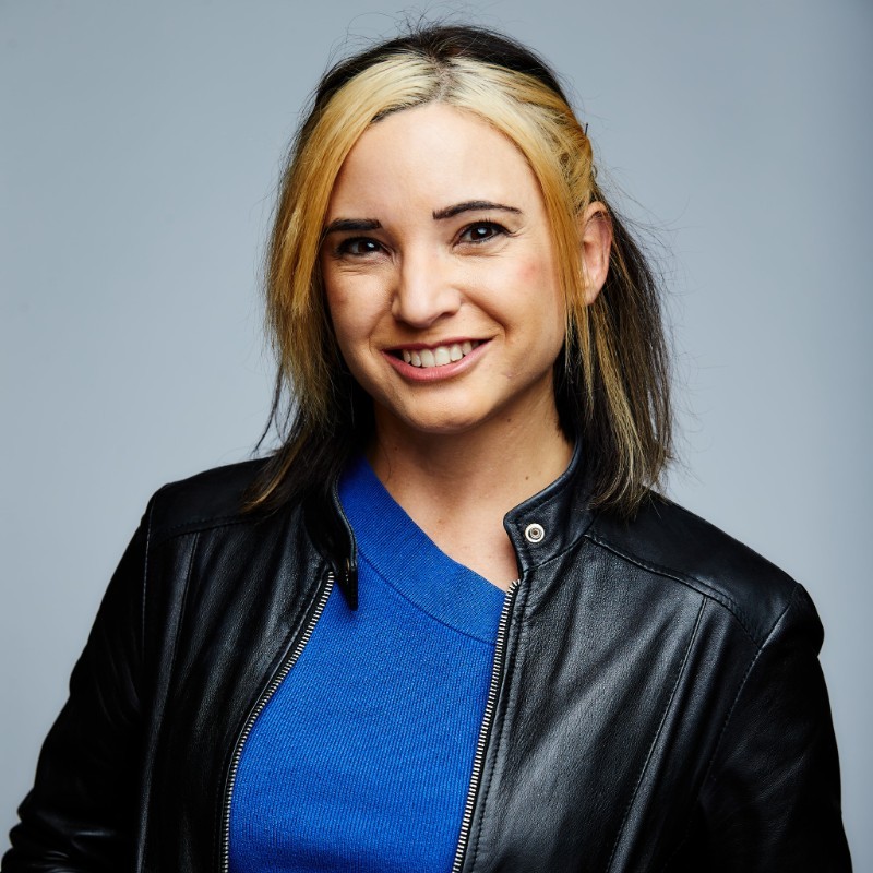 Smiling person with black and blonde hair wearing a blue shirt and black leather jacket, posed against a plain gray background.
