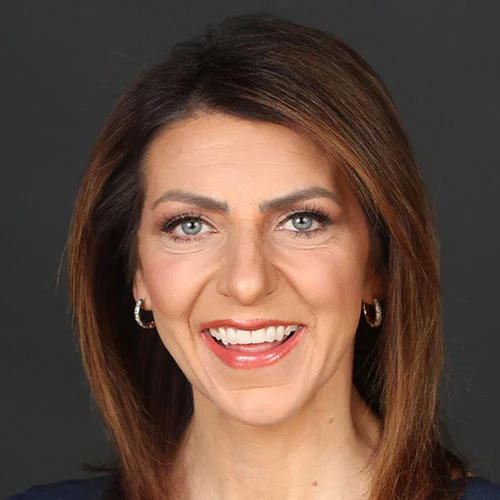 A woman with straight brown hair, blue eyes, and light makeup is smiling at the camera against a plain dark background. She is wearing hoop earrings.