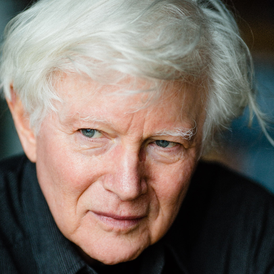 An older man with white hair and blue eyes looks slightly to the side, wearing a dark shirt, with a neutral expression.