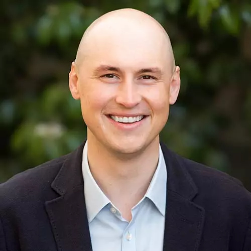 A smiling, bald man in a dark blazer and light blue shirt poses outdoors in front of blurred greenery.