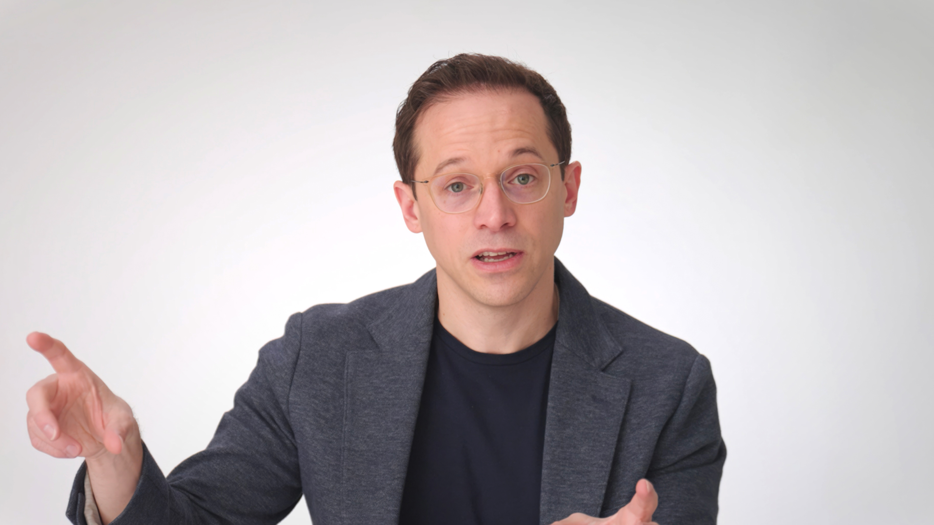 A man wearing glasses and a dark blazer gestures with his left hand while looking forward against a plain light background.