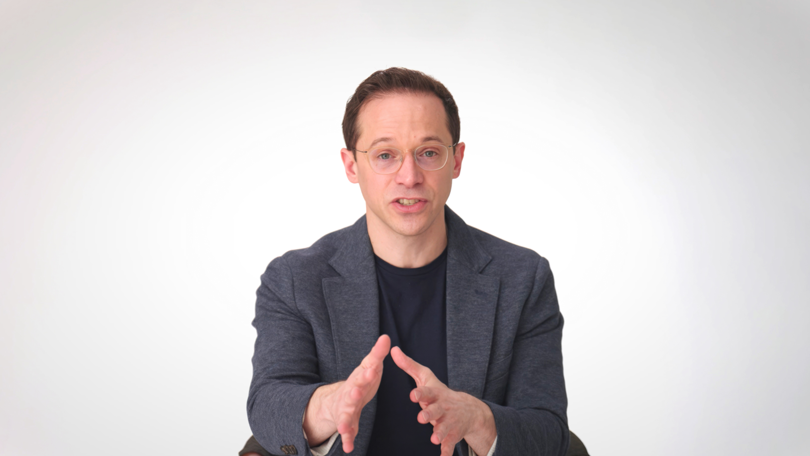 A man wearing glasses and a navy blazer speaks while gesturing with his hands against a plain white background.