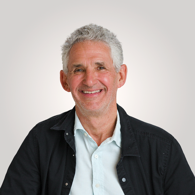 An older man with short gray hair and a light beard is smiling, wearing a light blue shirt and a black jacket, against a plain light background.