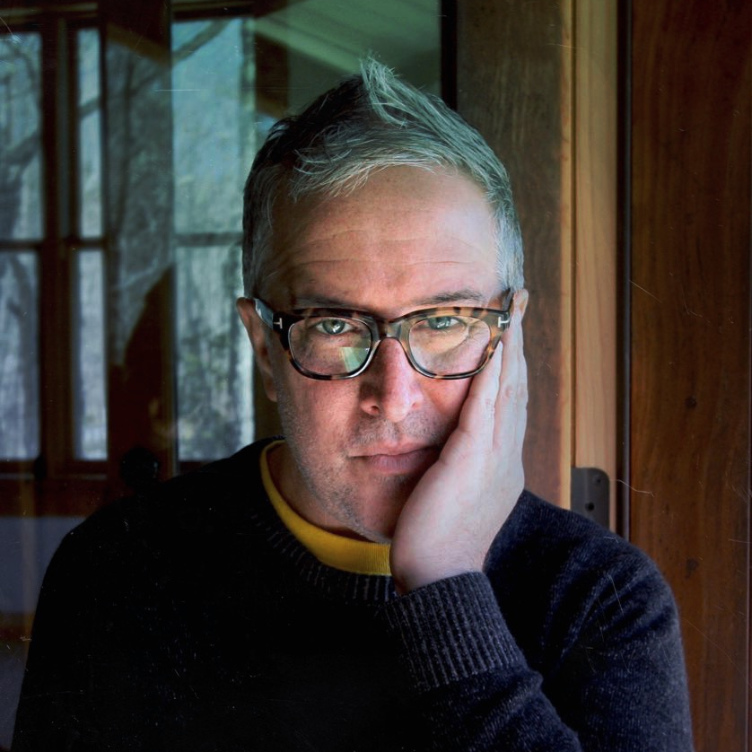 Middle-aged person with short gray hair and glasses rests their face on their hand while looking directly at the camera, indoors near a window.