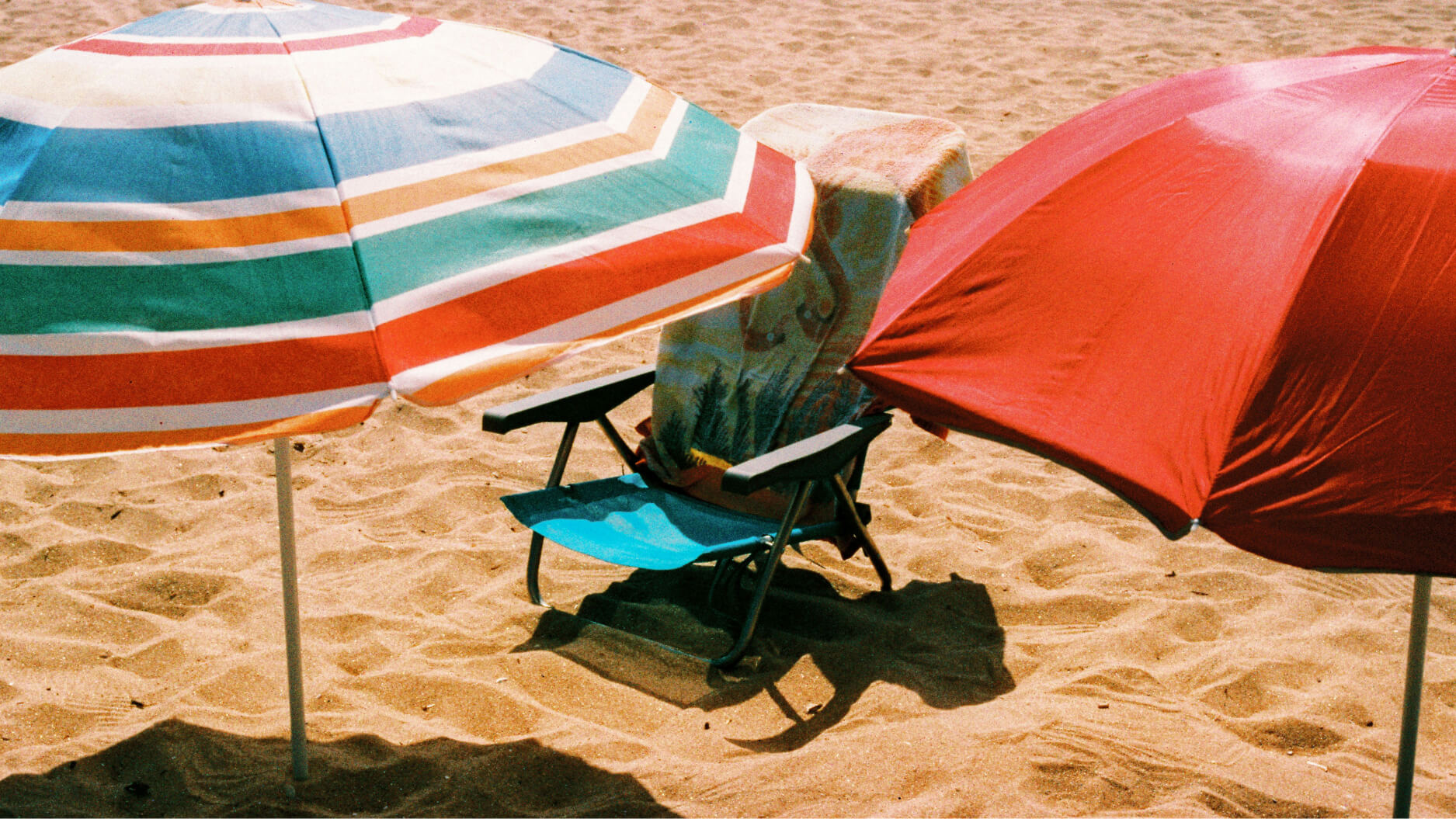 Two beach umbrellas, one striped and one red, shade a folding chair with a towel draped over it on the sandy beach—inviting you to rest and unwind by the shore.