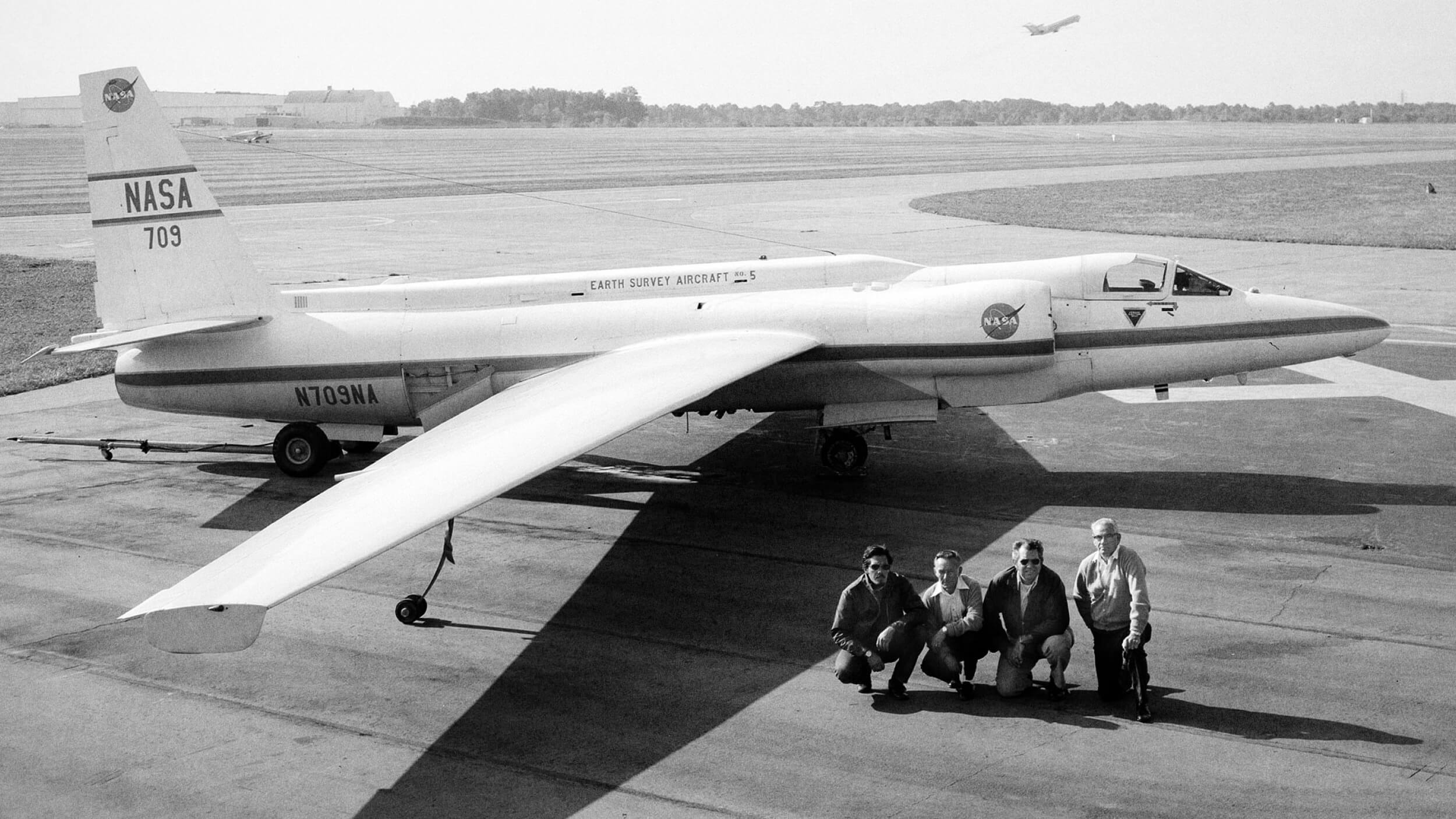 Black and white photo of a NASA Earth Survey Aircraft parked on a tarmac, with four people crouching in front of it and another plane taking off in the background.